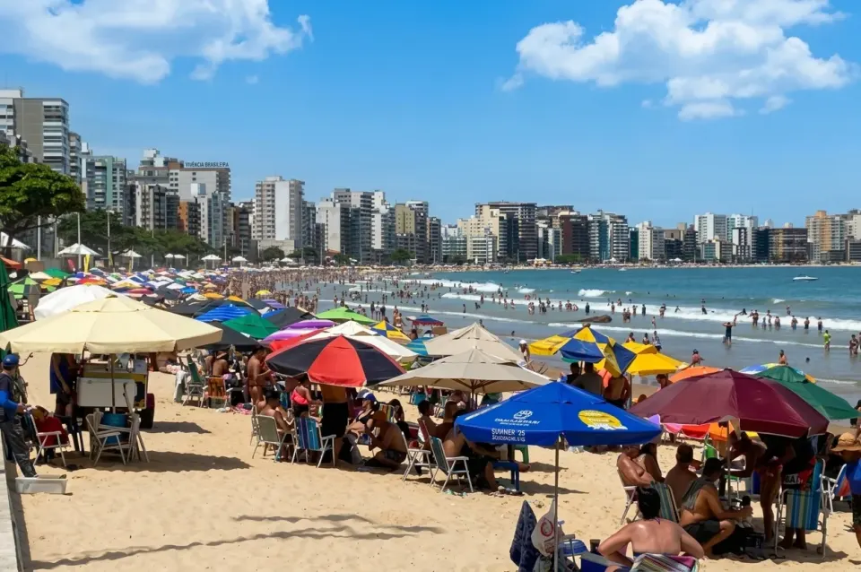 Praia do Morro cheia em dia de sol, com guarda-s&oacute;is coloridos na areia e os pr&eacute;dios da orla ao fundo, em Guarapari