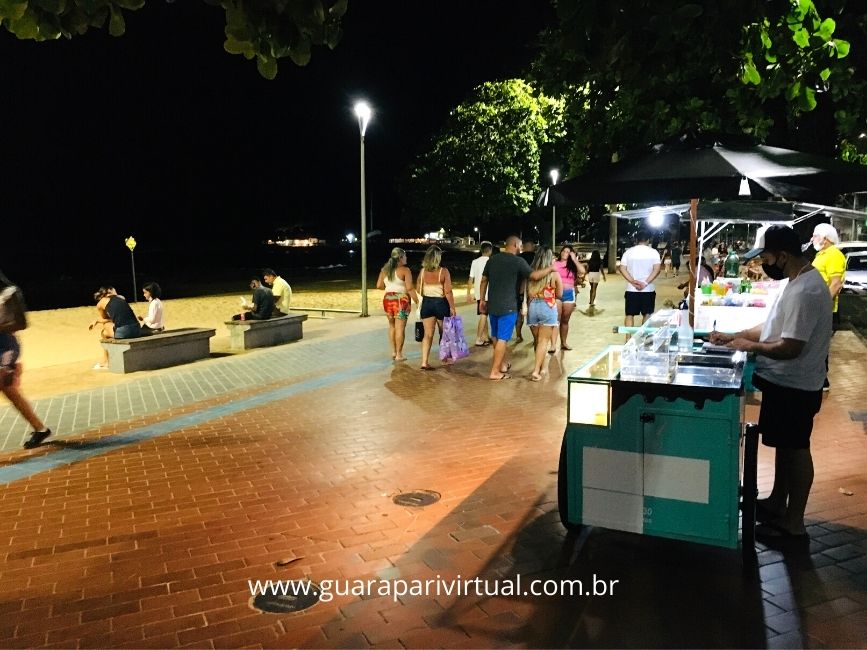 Orla iluminada do Centro de Guarapari &agrave; noite, com pessoas passeando no cal&ccedil;ad&atilde;o e um carrinho de lanches