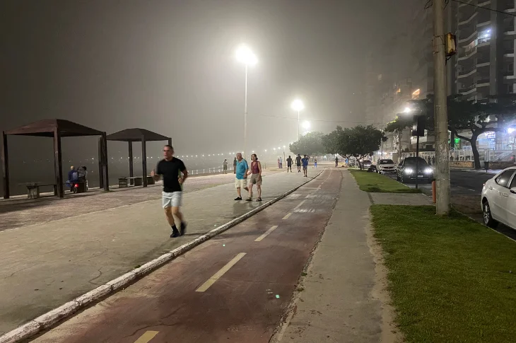 Cal&ccedil;ad&atilde;o e ciclovia da Praia do Morro &agrave; noite, iluminados, com pessoas caminhando e neblina sobre a orla, em Guarapari