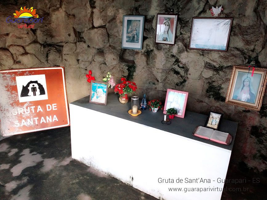 Interior da Gruta de Sant'Ana, com altar branco, flores, velas e quadros de santas nas paredes de pedra