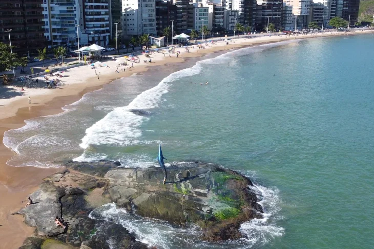 Vista do alto da Praia do Morro, em Guarapari, com a est&aacute;tua do Marlim Azul sobre as pedras e a orla de pr&eacute;dios ao fundo