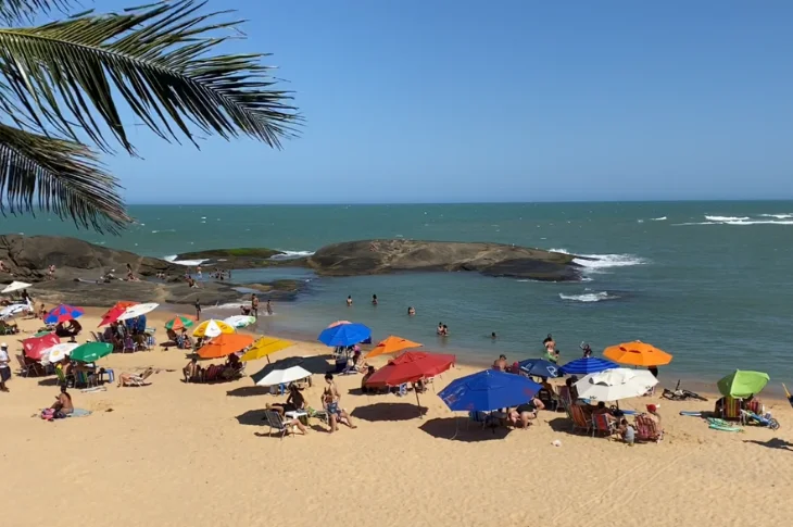 Banhistas e guarda-s&oacute;is coloridos na Praia dos Namorados, com pedras no mar e coqueiro em primeiro plano, em Guarapari