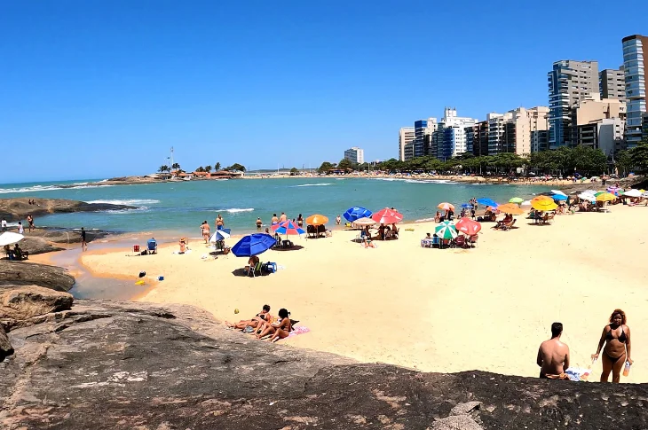 Praia dos Namorados, no Centro de Guarapari, com areia dourada, pedras e pr&eacute;dios ao fundo