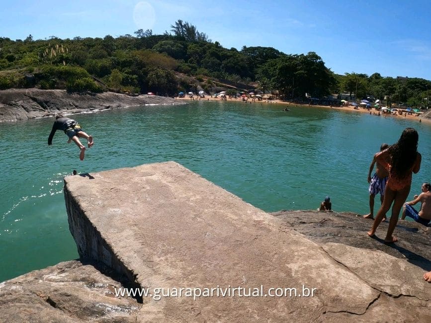 Jovem saltando da Pedra do Mero no mar verde, com a praia e a vegeta&ccedil;&atilde;o de Setiba ao fundo, em Guarapari