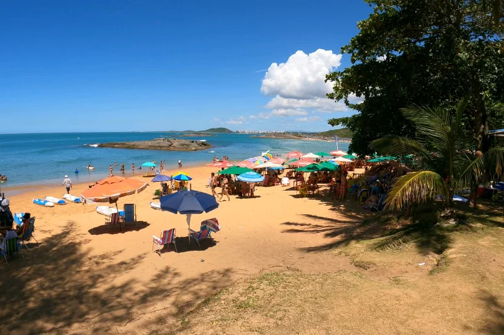 Orla da Praia de Setiba com guarda-s&oacute;is coloridos, mar calmo e a ilhota de pedra ao fundo, em Guarapari